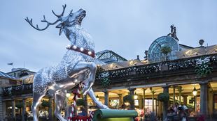 Статуя белого оленя, украшенная рождественскими огнями, стоит напротив освещенного здания рынка Ковент-Гарден.