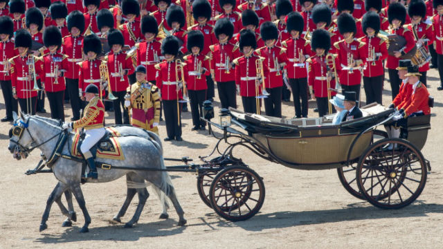 Horses pulling a vehicle in which the Queen is sitting, with guards wearing red uniforms in the background.