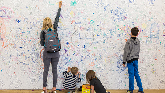 Дама и трое детей рисуют на белой стене, на которой уже много рисунков.