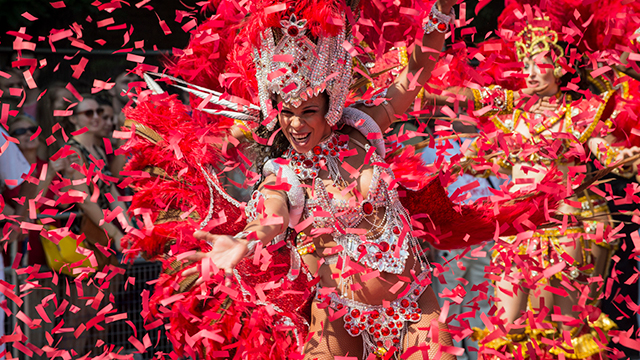 Дама, одетая в карнавальный головной убор и одежду, танцует, пока красный тикер наполняет воздух.