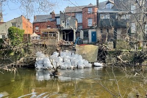 Белые мешки с камнями на берегу реки перед несколькими домами.